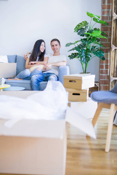 Young beautiful couple sitting on the sofa drinking coffee at new home around cardboard boxes - Foto, Imagem