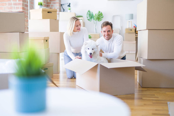 Young beautiful couple with dog sitting on the floor at new home around cardboard boxes - Foto, Bild