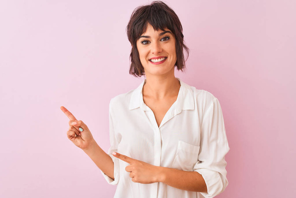 İzole edilmiş pembe arka planda duran beyaz gömlekli genç bir kadın gülümsüyor ve kameraya bakıyor iki eliyle ve parmaklarıyla kenarı işaret ediyor.. - Fotoğraf, Görsel
