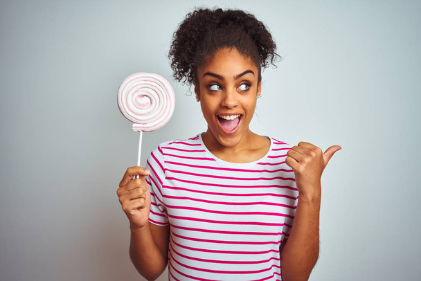 Африканська американка - підліток, яка їсть барвисті цукерки над ізольованим білим фоном і показує пальцем до сторони з радісним обличчям посміхаючись - Фото, зображення
