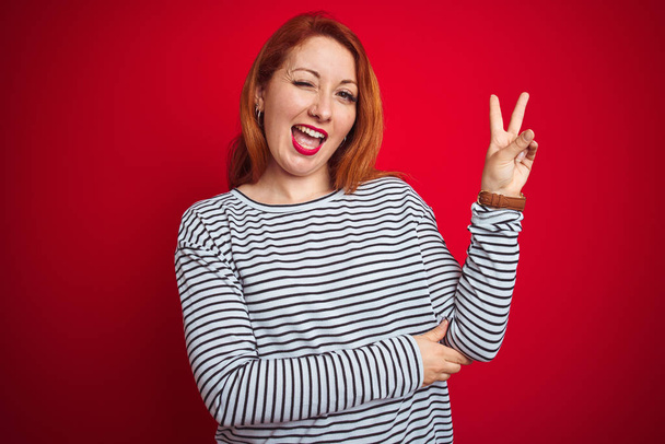 Jeune femme rousse portant des bretelles chemise marine debout sur fond rouge isolé souriant avec un sourire heureux clin d'oeil à la caméra faisant signe de victoire. Numéro deux
. - Photo, image