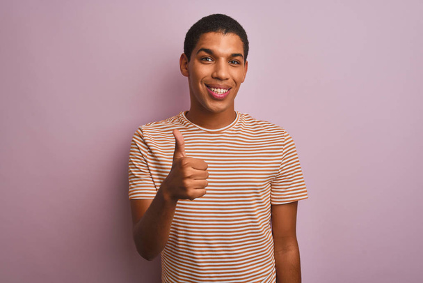 junger gutaussehender arabischer Mann in gestreiftem T-Shirt, der vor isoliertem rosa Hintergrund steht und mit der Hand fröhlich Daumen nach oben gestikuliert. zustimmender Ausdruck, der mit Erfolg in die Kamera blickt. - Foto, Bild
