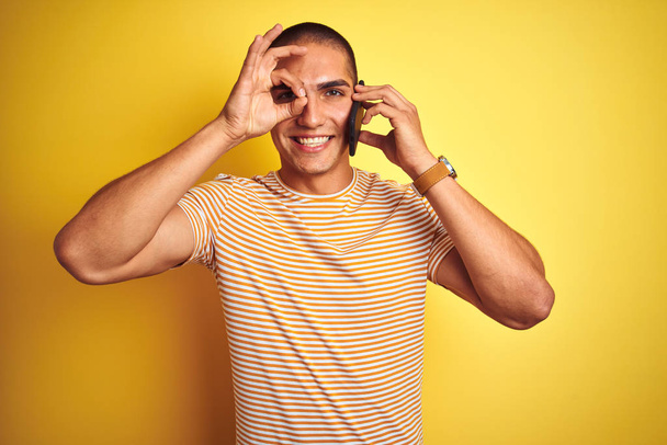 Молодой красивый мужчина говорит с помощью смартфона на желтом изолированном фоне со счастливым лицом улыбается делает хорошо знак с рукой на глазу глядя сквозь пальцы
 - Фото, изображение