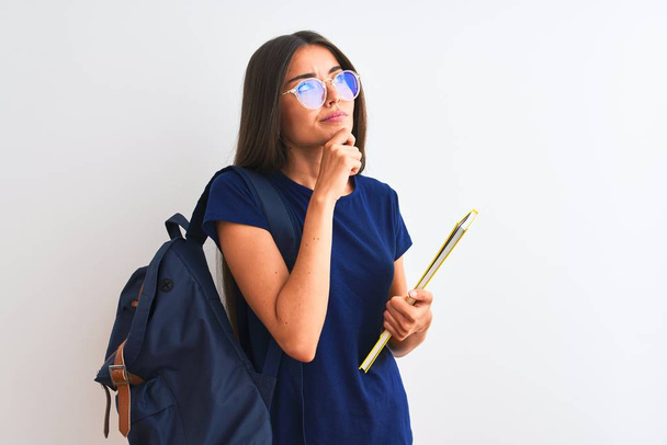 Młoda studentka nosząca okulary z plecakiem trzymająca książkę na białym tle poważna twarz myśląca o pytaniu, bardzo zdezorientowany pomysł - Zdjęcie, obraz