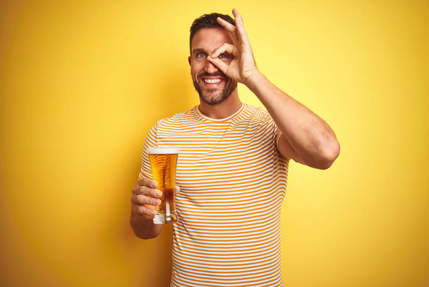 若いですハンサムな男飲むA pintガラスのビール上に隔離された黄色の背景とともに幸せな顔笑顔doing ok signとともに手で目を通して指 - 写真・画像