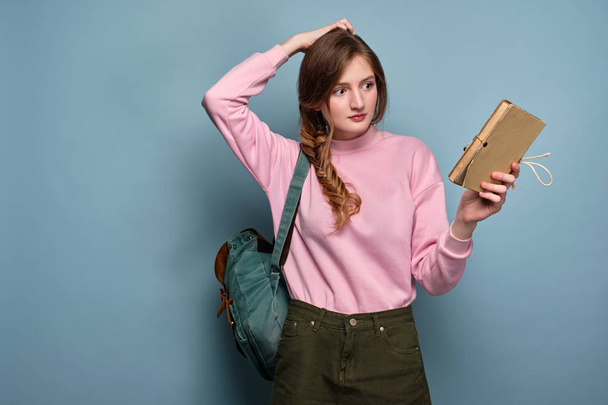 A girl in a pink sweater with a backpack stands on a blue background and holds a book, looking puzzled at her, scratching her head - Fotó, kép