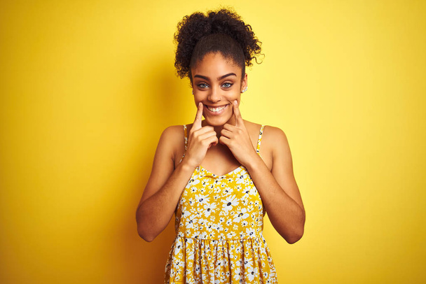 Femme afro-américaine portant une robe florale décontractée debout sur un fond jaune isolé Souriant avec la bouche ouverte, les doigts pointant et forçant sourire joyeux
 - Photo, image