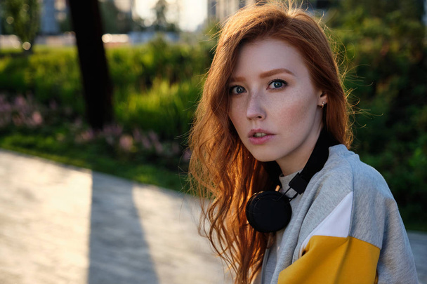 Portrait of a red-haired girl with freckles and blue eyes sitting in a park with headphones on her neck in the sunset rays - Fotó, kép