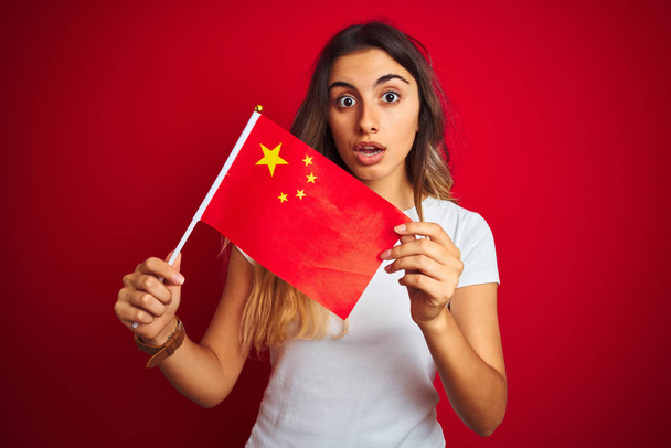 Fiatal nő tartja zászló Kína felett piros elszigetelt háttér megijedt a sokk egy meglepetés arc, fél, és izgatott a félelem kifejezés - Fotó, kép