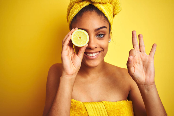 Αφρογυναίκα φορώντας πετσέτα μετά το ντους κρατώντας φέτα λεμόνι πάνω από απομονωμένο κίτρινο φόντο κάνει εντάξει σημάδι με τα δάχτυλα, εξαιρετικό σύμβολο - Φωτογραφία, εικόνα