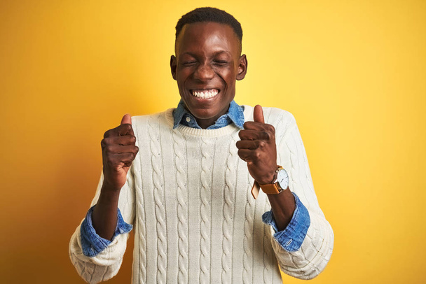 Африканський американець, одягнений в джинсову сорочку і білий светр над жовтим фоном, схвильований успіхом з піднятими руками, закрив очі, щоб відсвяткувати перемогу, посміхаючись. Концепція переможця. - Фото, зображення