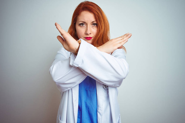 Молода руда жінка-лікар, використовуючи стетоскоп над білим ізольованим тлом Вираз відторгнення схрещування рук робить негативний знак, сердите обличчя
 - Фото, зображення