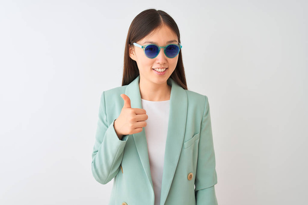 Empresaria china con chaqueta y gafas de sol sobre fondo blanco aislado feliz con una gran sonrisa haciendo signo ok, pulgar hacia arriba con los dedos, señal excelente
 - Foto, Imagen