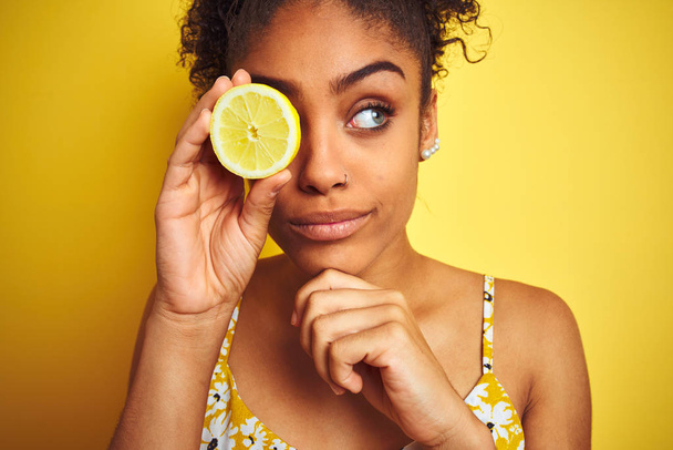 Fiatal afro-amerikai nő kezében szelet citrom felett elszigetelt sárga háttér komoly arc gondolkodás kérdés, nagyon zavaros ötlet - Fotó, kép