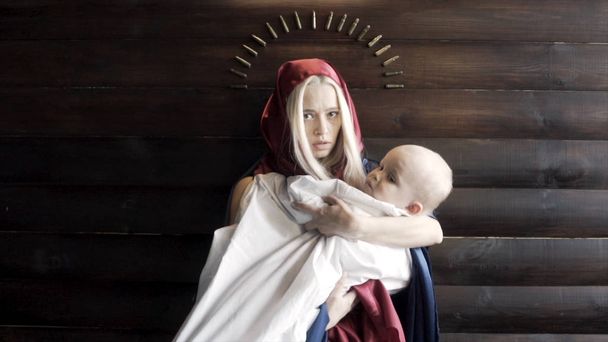 Woman with the covered head, mother with a baby in her arms standing against wooden wall with an arc of bullets, religion and war concept. Footage. The image of the virgin Mary and a Jesus. - Photo, Image