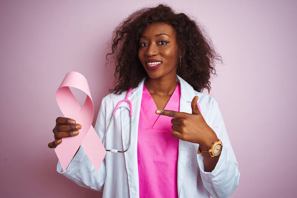 Молодая африканская американка доктор женщина с раковой лентой на изолированном розовом фоне очень счастлива указывая рукой и пальцем
 - Фото, изображение