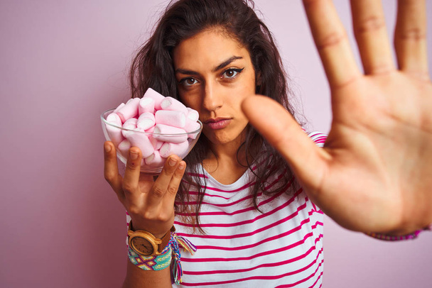 Молодая красивая женщина держит миску с зефиром на изолированном розовом фоне с открытой рукой делает знак стоп с серьезным и уверенным выражением, защитный жест
 - Фото, изображение