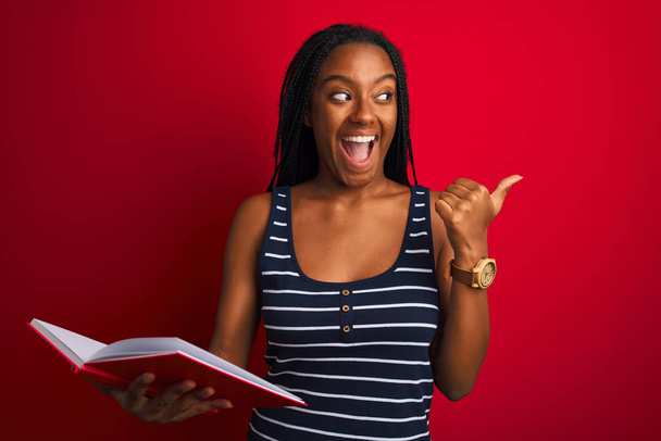 junge afrikanisch-amerikanische Studentin liest Buch steht über isolierten roten Hintergrund zeigt und zeigt mit dem Daumen zur Seite mit glücklichem Gesicht lächelnd - Foto, Bild