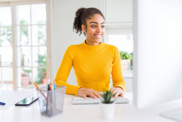若いアフリカ系アメリカ人の女の子がコンピュータを使って仕事をしていて、笑顔で自信を持って歯を見せています - 写真・画像