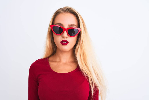 Jonge mooie vrouw met rood t-shirt en zonnebril over geïsoleerde witte achtergrond met serieuze uitdrukking op gezicht. Eenvoudig en natuurlijk kijkend naar de camera. - Foto, afbeelding