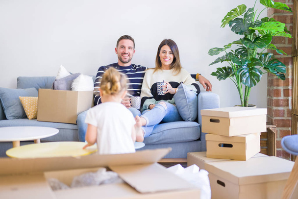 Hermosa familia, padres sentados en el sofá bebiendo café mirando a su hijo jugando en un nuevo hogar alrededor de cajas de cartón
 - Foto, imagen