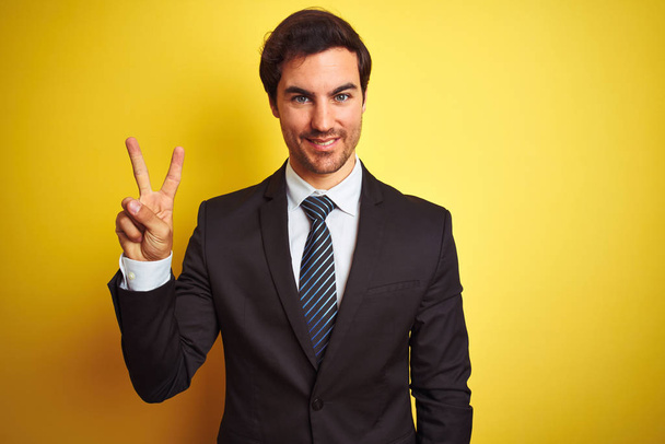 Młody przystojny biznesmen w garniturze i krawacie stojący nad odizolowanym żółtym tłem pokazując i wskazując palcami numer dwa uśmiechając się pewny siebie i szczęśliwy. - Zdjęcie, obraz