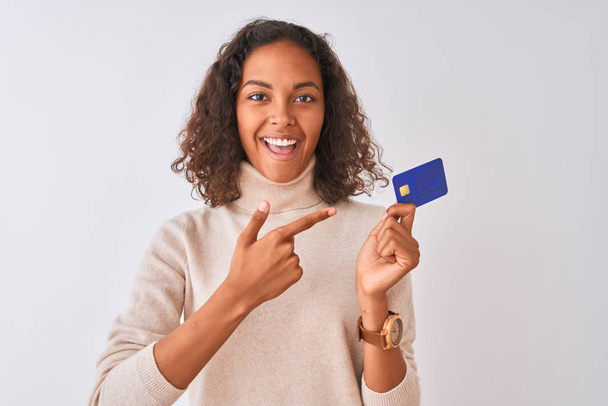 junge Brasilianerin mit Kreditkarte vor isoliertem weißem Hintergrund sehr glücklich mit Hand und Finger zeigend - Foto, Bild