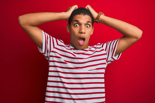 Young handsome arab man wearing striped t-shirt over isolated red background Crazy and scared with hands on head, afraid and surprised of shock with open mouth - Фото, зображення