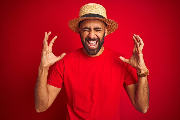Young handsome indian man wearing t-shirt and hat over isolated red background celebrating mad and crazy for success with arms raised and closed eyes screaming excited. Winner concept - Photo, Image