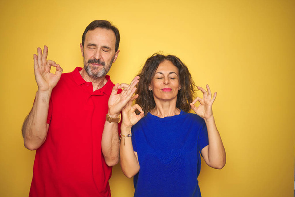 Beautiful middle age couple together standing over isolated yellow background relax and smiling with eyes closed doing meditation gesture with fingers. Yoga concept. - Photo, Image