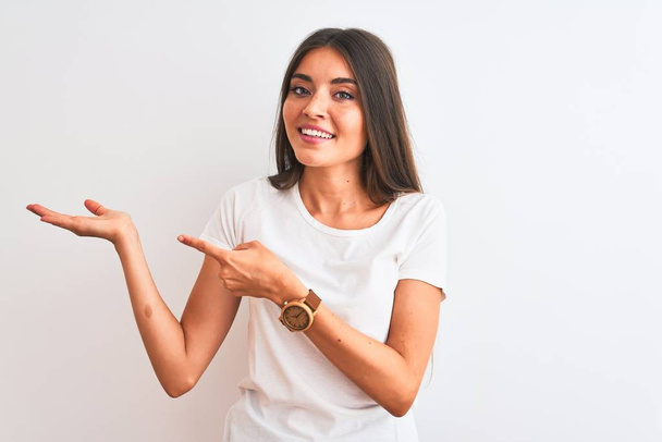 Young beautiful woman wearing casual t-shirt standing over isolated white background amazed and smiling to the camera while presenting with hand and pointing with finger. - Φωτογραφία, εικόνα