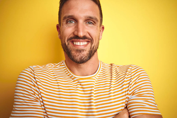 Detailní záběr mladého pohledného muže v neformálním tričku přes žluté izolované pozadí šťastný obličej s úsměvem se zkříženýma rukama dívá do kamery. Pozitivní osoba. - Fotografie, Obrázek