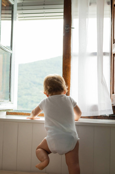 glückliches blondes Baby, das auf dem Bett sitzt, spielt und zu Hause ins Fenster schaut - Foto, Bild