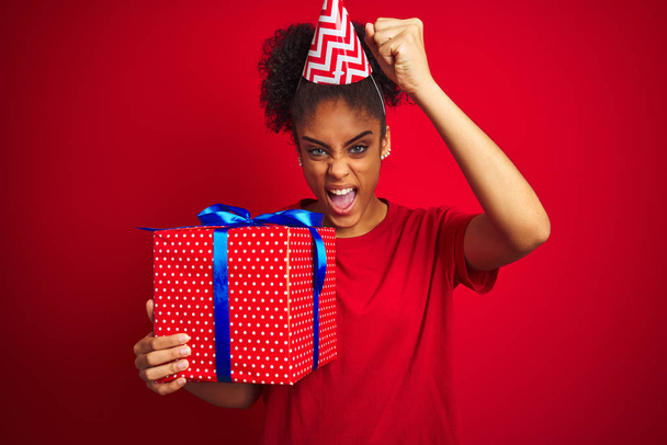Komik şapkalı Afrikalı bir kadın izole edilmiş kırmızı arka planda doğum günü hediyesi tutuyor. Sinirli ve sinirli bir şekilde bağırıyor, çıldırıyor ve el kaldırarak bağırıyor. Öfke kavramı. - Fotoğraf, Görsel