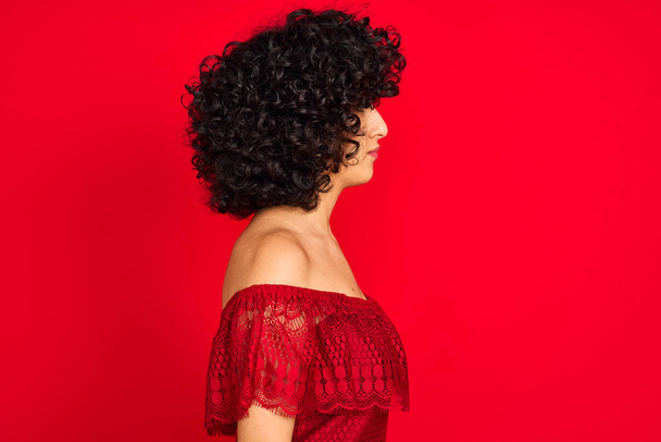 Jeune femme arabe aux cheveux bouclés portant une robe décontractée sur fond rouge isolé regardant vers le côté, pose de profil relax avec un visage naturel avec un sourire confiant
. - Photo, image