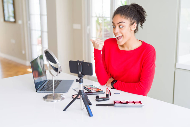 Jonge Afrikaanse Amerikaanse influencer vrouw opnemen make-up tutorial glimlachen met gelukkig gezicht kijken en wijzen naar de zijkant met duim omhoog. - Foto, afbeelding