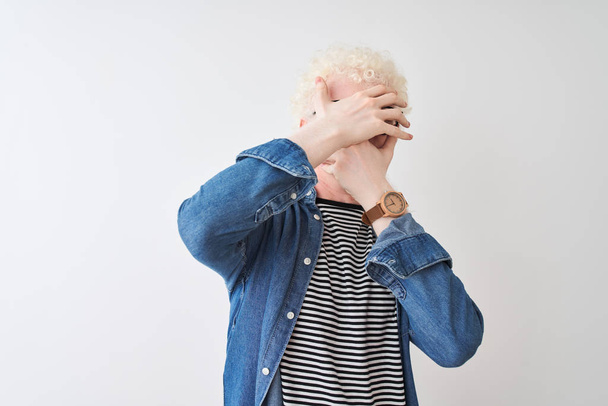 Junge Albino-Blonde mit Jeanshemd und Brille vor isoliertem weißen Hintergrund, die Augen und Mund mit Händen bedeckt, überrascht und schockiert. Emotionen verbergen - Foto, Bild