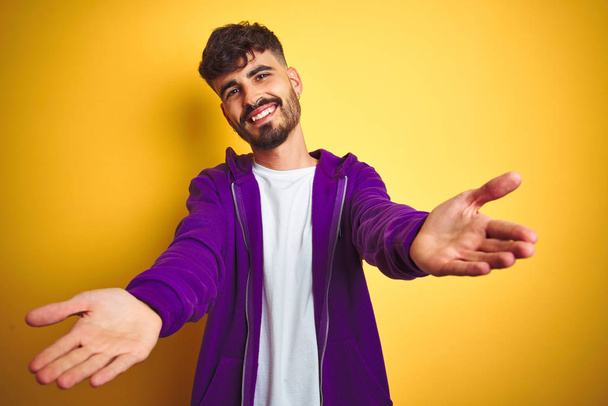 独立した黄色の背景の上にスポーツ紫のスウェットシャツを着てタトゥーを持つ若い男は抱擁のためのオープン腕で笑顔カメラを見て。幸せを受け入れる陽気な表情. - 写真・画像