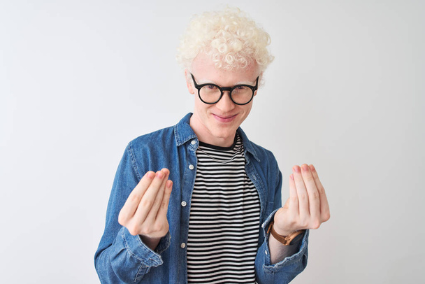 Jonge albino blonde man draagt denim shirt en bril over geïsoleerde witte achtergrond doen geld gebaar met de handen, vragen om salaris betaling, miljonair bedrijf - Foto, afbeelding