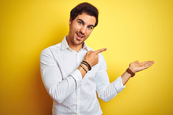 Young handsome businessman wearing elegant shirt standing over isolated yellow background amazed and smiling to the camera while presenting with hand and pointing with finger. - Φωτογραφία, εικόνα