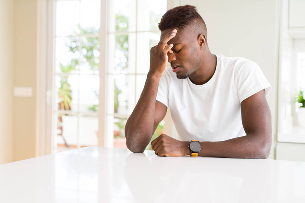 Beyaz masadaki yakışıklı Afro-Amerikalı adam yorgun, burnunu ve gözlerini ovuşturup yorgun ve baş ağrısı çekiyor. Stres ve hüsran kavramı. - Fotoğraf, Görsel