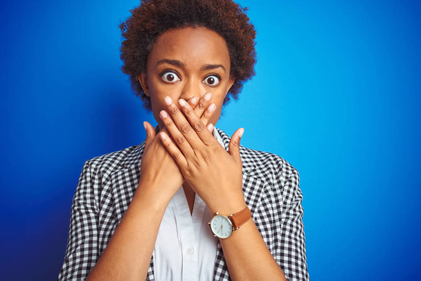 孤立した青い背景の上のアフリカ系アメリカ人のビジネスエグゼクティブ女性は、間違って手で口を覆ってショックを受けました。秘密の概念. - 写真・画像