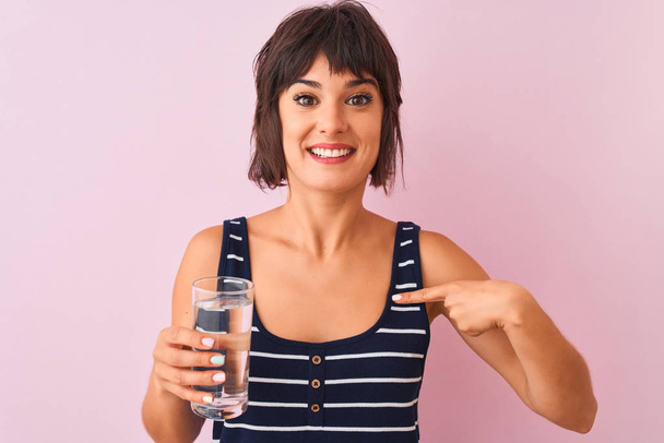 Jovem bela mulher segurando vidro de água em pé sobre fundo rosa isolado com rosto surpresa apontando dedo para si mesmo
 - Foto, Imagem