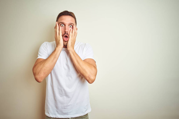 Young handsome man wearing casual white t-shirt over isolated background afraid and shocked, surprise and amazed expression with hands on face - 写真・画像
