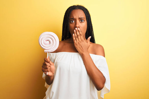 Genç Afro-Amerikalı kadın, izole edilmiş sarı arka planda duran şeker yiyor. Ağzı kapalı, utancından ağzı açık, korkusunun dışa vurumu, sessizlikten korkması, gizli konsept. - Fotoğraf, Görsel