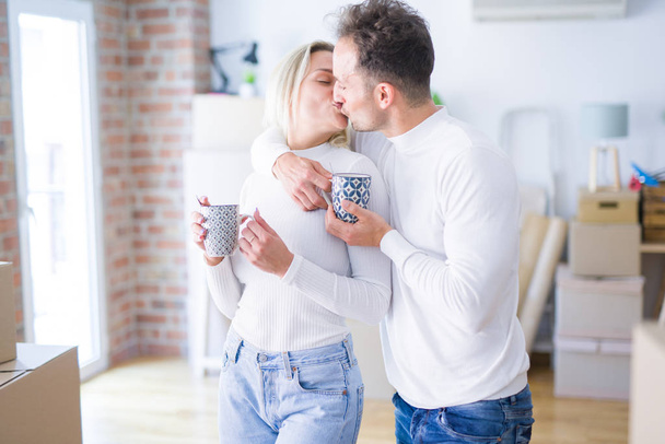 Jeune beau couple debout boire une tasse de café et embrasser à la nouvelle maison autour des boîtes en carton
 - Photo, image