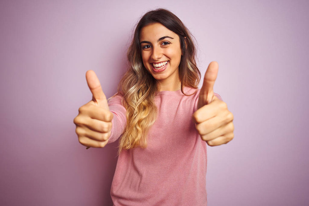 junge schöne Frau trägt einen Pullover über rosa isolierten Hintergrund Erfolgszeichen tun positive Geste mit der Hand, Daumen nach oben lächelnd und glücklich. Fröhlicher Ausdruck und Siegergeste. - Foto, Bild