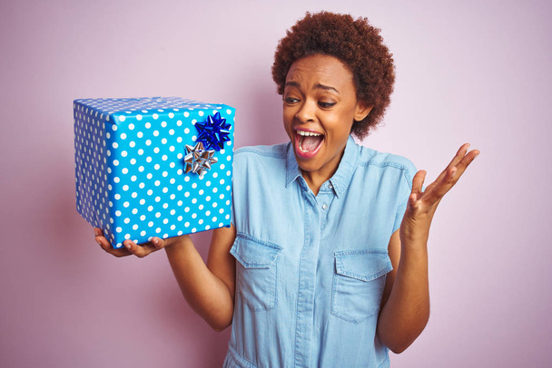 Afro-Amerikalı bir kadın pembe izole edilmiş arka planda doğum günü hediyesini tutarken mutlu ve heyecanlı, kazanan yüz ifadesi zaferi büyük bir gülümsemeyle kutluyor ve ellerini kaldırıyor. - Fotoğraf, Görsel
