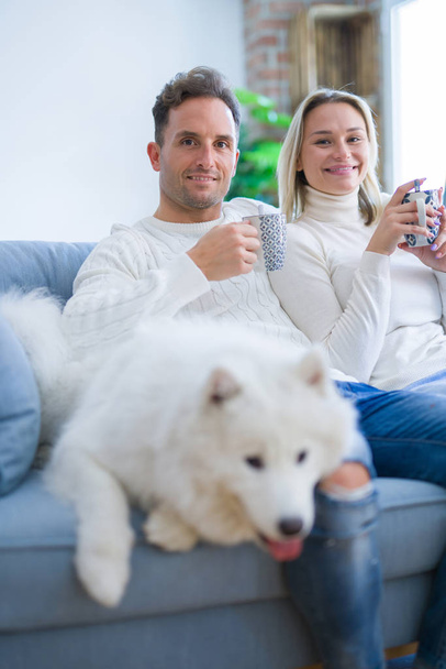 Молодая красивая пара с собакой, сидящей на диване, пьет кофе в новом доме вокруг картонных коробок
 - Фото, изображение