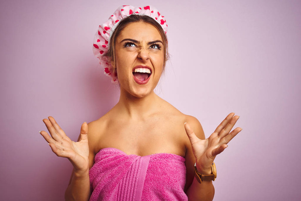 Mujer hermosa joven con toalla y gorra de ducha sobre fondo rosa aislado loco y loco gritando y gritando con expresión agresiva y los brazos levantados. Concepto de frustración
. - Foto, imagen